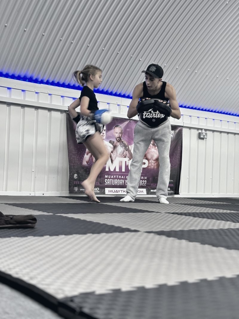 Jayden Gill holding a belly pad while a young student kicks it